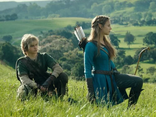 Dois personagens em trajes de fantasia, sentados em um campo verde amplo, observando a paisagem ao redor. A cena transmite clima de aventura e remete ao universo de Zelda.